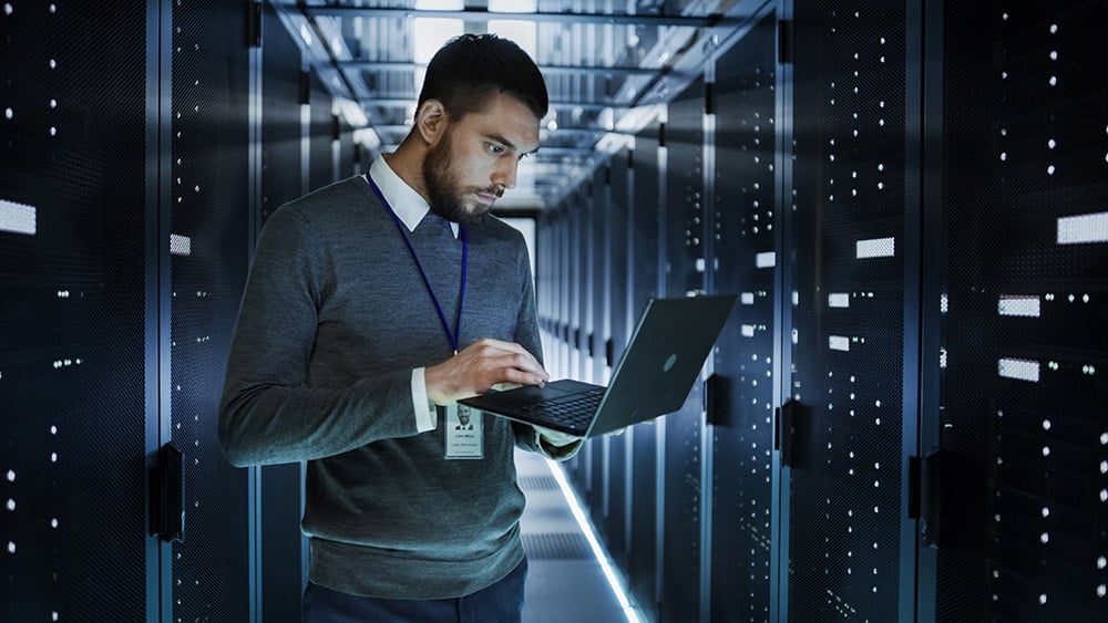 IT Technician Works on a Laptop in Big Data Center full of Rack Servers.